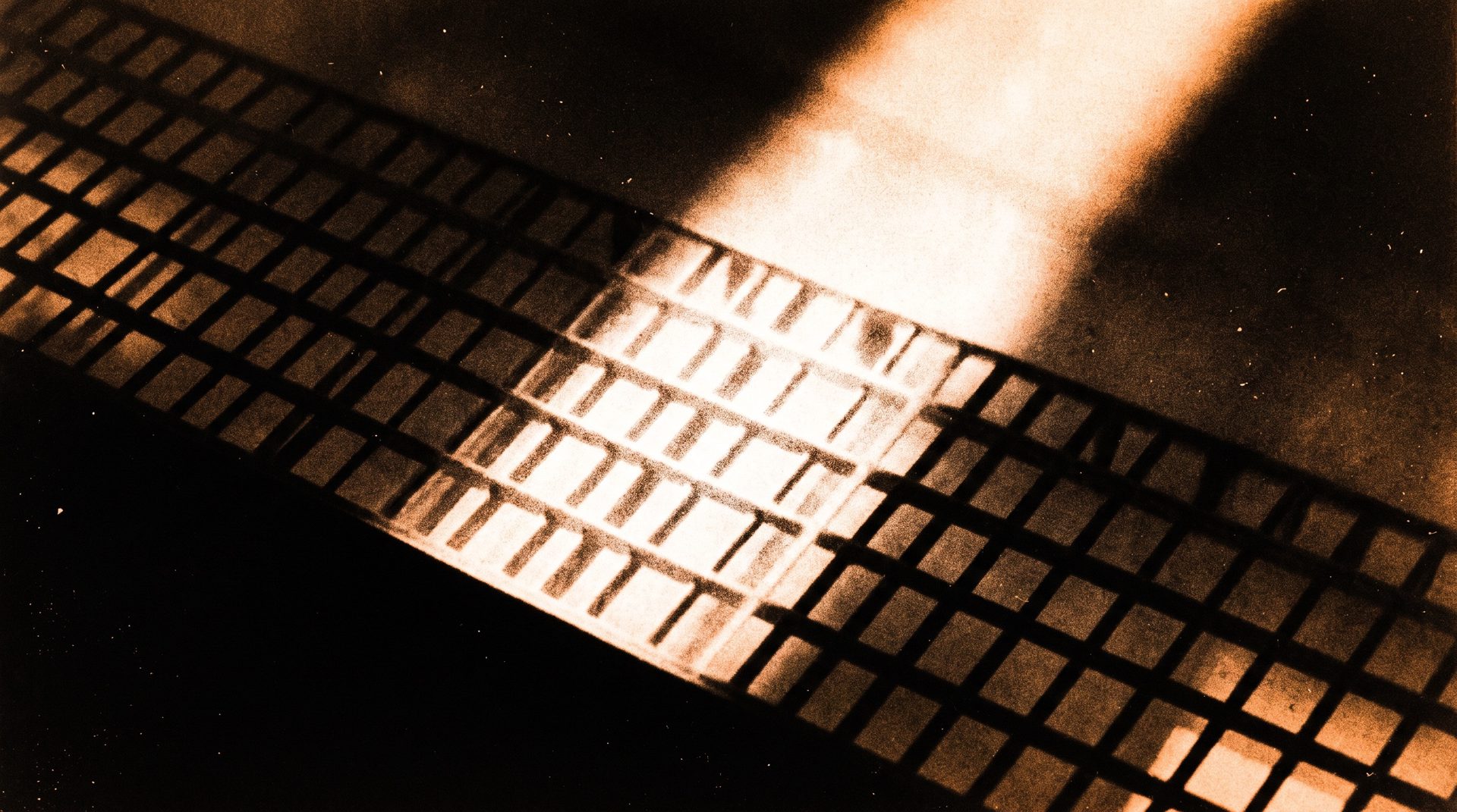 Photogram: the shadow lattice of a steel walkway grating seen from below, one broad shaft of light raking across the bars.